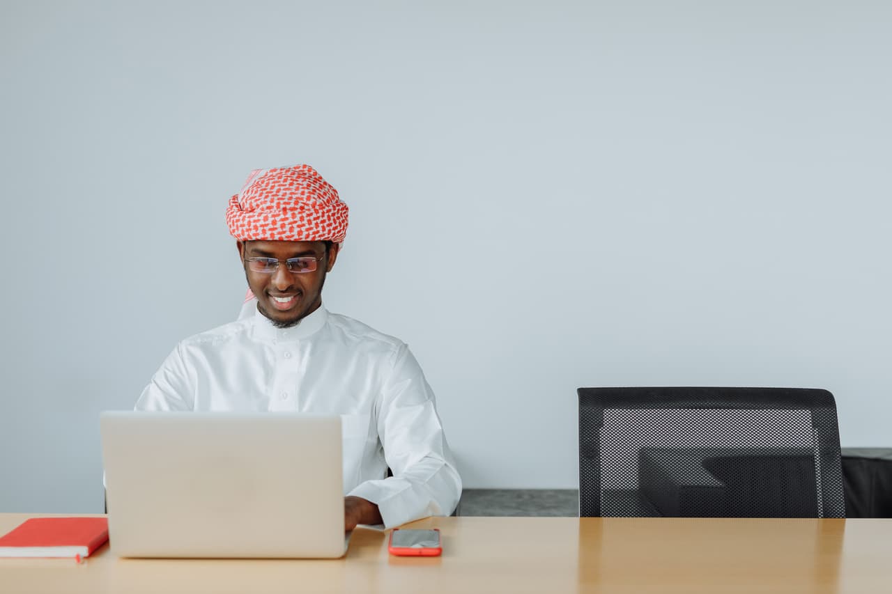 رجل يرتدي ثوبًا أبيض وشماغًا أحمر يبتسم وهو يعمل على حاسوب محمول في مكتبه